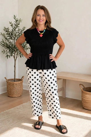 A woman modeling a black peplum top and white pants with black polka dots, standing in a bright room with a potted plant and a wooden bench in the background.
