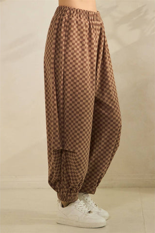 Brown and beige checkered harem pants worn by a model, paired with white sneakers, shown from the waist down against a neutral beige background.