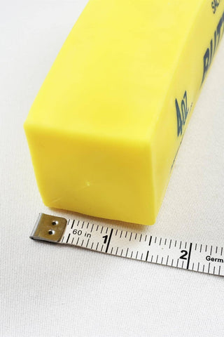 A yellow rectangular block of material, likely a sample of rubber or plastic, shown next to a metal tape measure on a white textured fabric background.