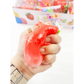 A hand holding a red squishy toy shaped like a strawberry, with a box of similar toys in the blurred background.