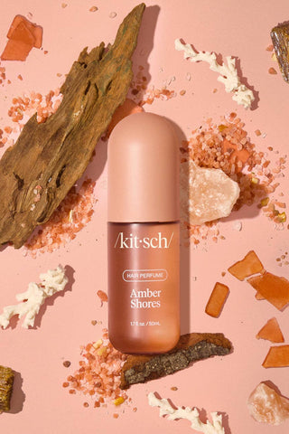 A bottle of Kit & Sch hair perfume labeled 'Amber Shores' is centered on a peach-pink background, surrounded by decorative natural elements including wood, salt crystals, and coral pieces. The product bottle is a warm peachy pink with amber-colored liquid visible through the lower portion.