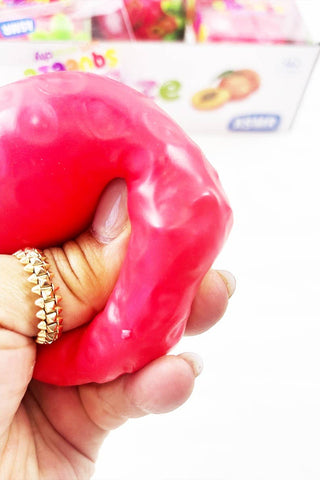 A hand squeezes a bright pink, bumpy, squishy toy against a white background, with a blurred box of similar toys visible in the background.