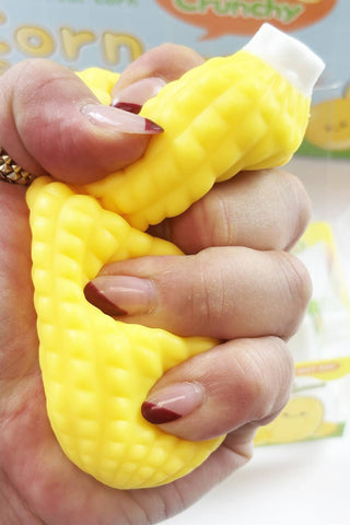 A hand squeezing a yellow corn-shaped squishy toy, with a blurred product box in the background.