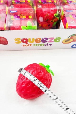 A red strawberry-shaped squeeze soft stretchy toy is shown in front of its branded packaging box, with a measuring tape wrapped around the toy to indicate size. The box reads 'squeeze soft stretchy' and shows fruit illustrations.