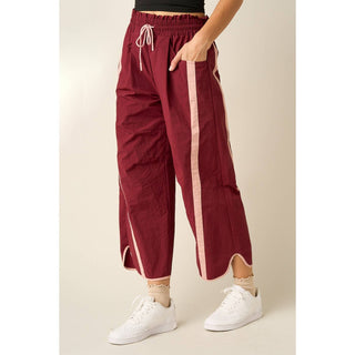 A person wearing loose burgundy cropped pants with light beige side stripes and white sneakers, standing against a plain beige studio background.