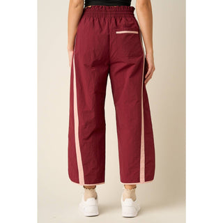 A person shown from the back wearing wide-leg cropped pants in burgundy with light pink side stripes and a matching back pocket detail, paired with white sneakers against a plain beige studio background.