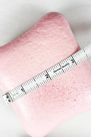 A close-up of a pink toast squishy with small darker pink speckles, shown with a white measuring tape laid across it on a light background.