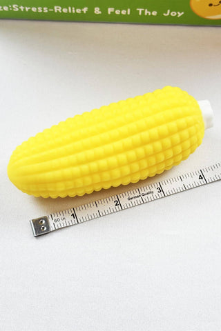 A bright yellow corn-shaped stress relief toy with a white handle, shown next to a metal measuring tape on a white surface.
