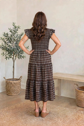 A woman stands with her back to the camera wearing a brown and black plaid tiered midi dress with ruffled cap sleeves. She is in a styled indoor setting with a potted olive tree, a wooden bench, and woven baskets.