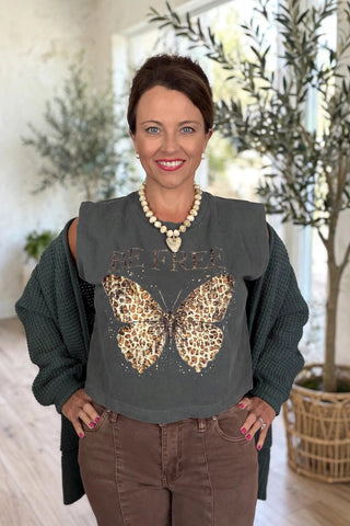 A woman wearing a dark gray sleeveless crop top with a leopard-print butterfly graphic and 'BE FREE' text, layered over a dark green knit sweater, brown pants, and a pearl necklace, standing in a brightly lit room with potted plants.
