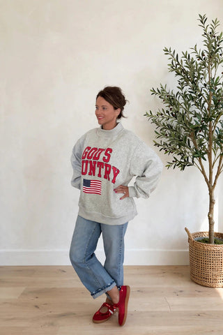 A woman modeling a light gray sweatshirt with 'GOD'S COUNTRY' and an American flag graphic in red, paired with blue jeans and red shoes, standing in a bright room next to a potted tree.