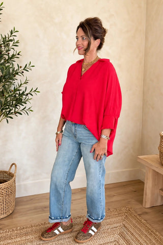 A woman stands in a bright room wearing a bright red draped long-sleeve top, light blue jeans, and red-and-white sneakers. A potted olive tree, a woven basket, and a wooden bench are visible in the background.