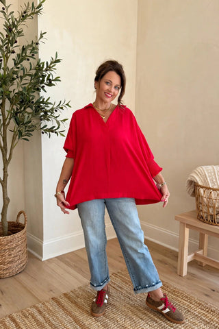 A woman modeling a bright red, loose-fitting short-sleeve top with light-wash blue jeans and brown slip-on shoes, standing in a brightly lit room with beige walls, a potted olive tree, and a wooden bench.