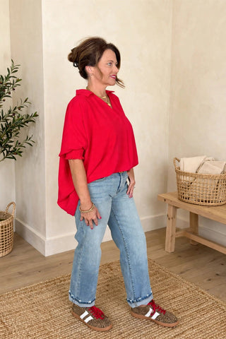 A woman wearing a bright red oversized short-sleeve top, light-wash blue jeans, and red-and-white striped sneakers stands in a styled room with a potted plant, woven bench, and rug.
