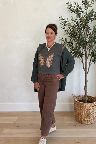 A woman modeling a dark olive green cardigan with a gold sequin butterfly design over a gray top, paired with brown corduroy pants and white sneakers, standing in a room with a potted olive tree.