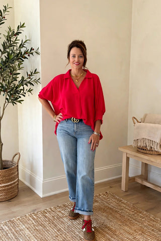 A woman stands in a bright, modern room wearing a vibrant red long-sleeve blouse, light-wash blue jeans, and red shoes. The room features a potted olive tree, a wooden bench with a throw blanket, and a woven rug.
