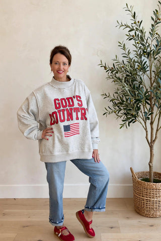 A woman stands indoors wearing a light heather gray sweatshirt with 'GOD'S COUNTRY' in red letters and a small American flag graphic, paired with light blue jeans and red Mary Jane shoes. She is posed next to a potted olive tree against a beige wall and light wood floor.