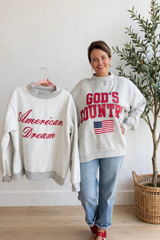 A woman stands indoors wearing a light gray sweatshirt with red 'God's Country' lettering and a small American flag graphic, while holding up a matching light gray sweatshirt on a hanger that reads 'American Dream' in red script. She is wearing blue jeans and red shoes, with a potted tree beside her in a bright room with a white wall and wood floor.