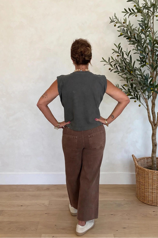 A woman stands with her back to the camera wearing a dark gray sleeveless top and brown wide-leg pants, posing in a room with a textured beige wall, light wood floor, and a potted olive tree.