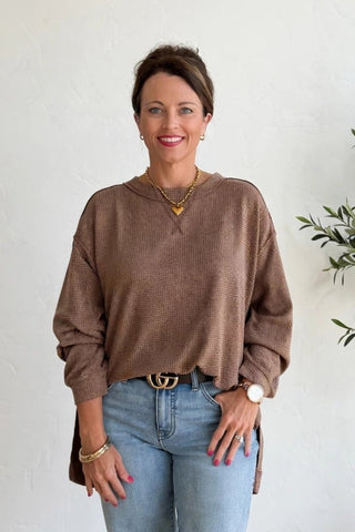 A woman stands in a bright room wearing a brown long-sleeve knit sweater, light blue jeans, and black slide sandals. She is positioned next to a potted olive tree against a white wall.