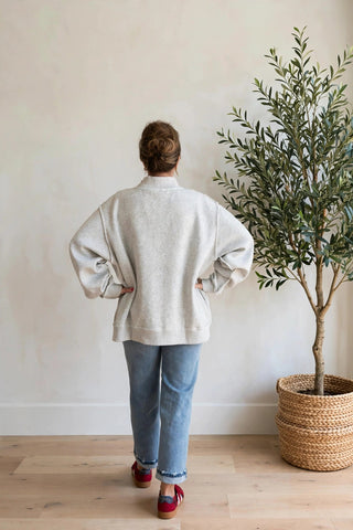 A person viewed from behind wearing a cream-colored knit sweater, light blue jeans, and red-and-white espadrille sandals, standing in a room with a light-colored wall, wooden floor, and a potted olive tree.