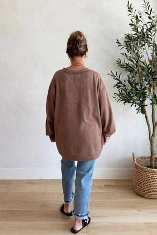 A woman viewed from behind wearing an oversized brown long-sleeve top, light blue jeans, and black slide sandals, standing in a room with a light wood floor, white wall, and a potted olive tree.