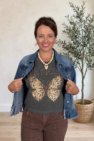 A woman modeling a dark gray graphic t-shirt with a large leopard-print butterfly design, styled with an open blue denim jacket, brown pants, and a pearl necklace in a bright indoor setting.