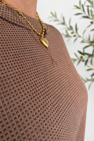 Close-up of a person wearing a brown waffle-knit top and a gold heart pendant necklace, with blurred green leaves in the background.