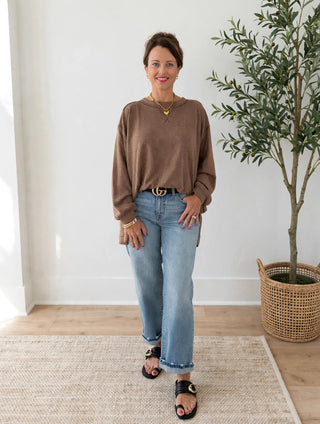 A woman stands indoors wearing a brown long-sleeve knit top, light blue jeans, and black slide sandals, with a gold belt and necklace. She is posed in a bright room with a potted olive tree and a woven rug.
