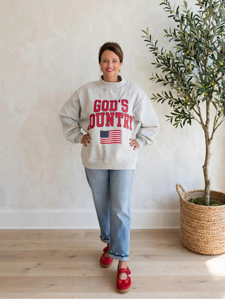 A woman stands indoors wearing a light gray sweatshirt with 'GOD'S COUNTRY' and an American flag graphic in red, paired with light blue jeans and red shoes. She is posed next to a tall potted olive tree against a textured off-white wall.