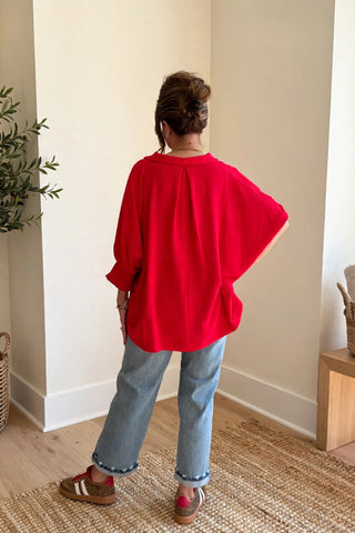 A woman stands with her back to the camera in a bright, modern room, wearing a vibrant red oversized short-sleeve top, light blue jeans, and brown sneakers.