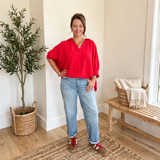 A woman stands indoors modeling a bright red loose blouse with light blue jeans and red-and-white sneakers. The room has cream walls, a potted tree, a woven bench with a throw, and a rug on a wood floor.