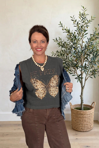 A woman wearing a dark gray sleeveless top with a leopard-print butterfly graphic and the word 'BE FREE across the chest, styled with a denim jacket draped off her shoulders and brown pants, standing in a room next to a potted olive tree.
