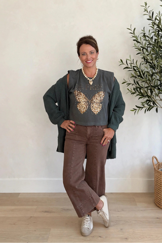 A woman posing in a room with a beige wall and wooden floor, wearing a dark green long-sleeve top with a gold butterfly graphic, brown wide-leg pants, and white sneakers.