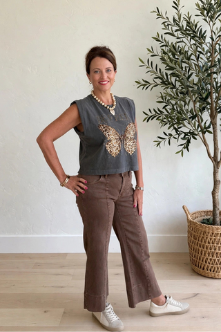 A woman posing in a room with a light-colored wall and wooden floor, wearing a dark gray sleeveless top with a gold butterfly design and brown wide-leg pants, standing next to a potted olive tree.