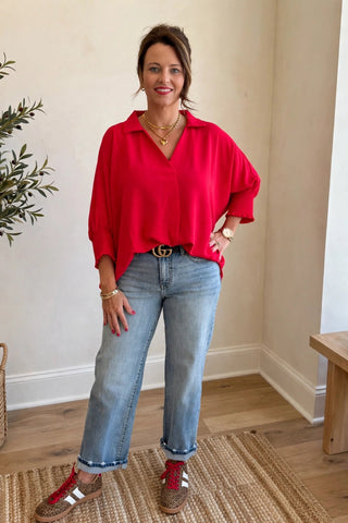 A woman stands indoors wearing a bright red oversized collared blouse, light blue jeans, red and white sneakers, and gold jewelry. She is posed in a room with a potted tree, woven baskets, a wooden bench, and a window.