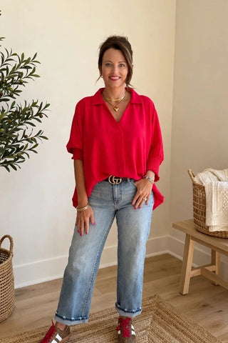 A woman stands in a bright, modern room wearing a vibrant red button-up shirt, light-wash blue jeans, and red-and-white sneakers. She is positioned in front of a beige wall with a potted plant on the left and a wooden bench with a basket on the right.
