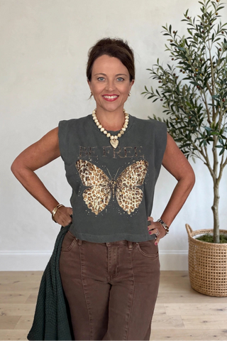 A woman posing indoors wearing a dark gray sleeveless top with a leopard-print butterfly graphic and the word 'BELIEVE' across the chest, paired with brown pants.