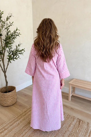 A woman viewed from behind wearing a pink and white gingham long robe in a bright, minimalist room with a potted plant and a wooden bench.