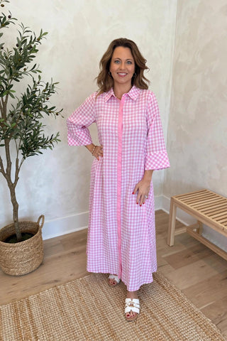 A woman modeling a pink and white gingham long-sleeve maxi shirt dress with a pink button-down front, standing in a brightly lit room with a potted olive tree and a wooden bench.