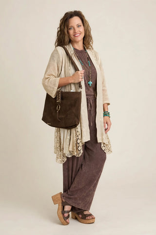 A woman modeling a bohemian-style outfit featuring a cream lace-trimmed cardigan over a brown tunic and wide-leg pants, carrying a brown suede shoulder bag against a warm beige studio background.