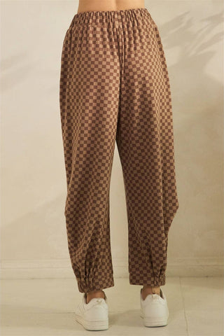 Brown and tan checkered harem pants with an elastic waistband and ankle cuffs, shown on a model wearing white sneakers against a neutral beige background.