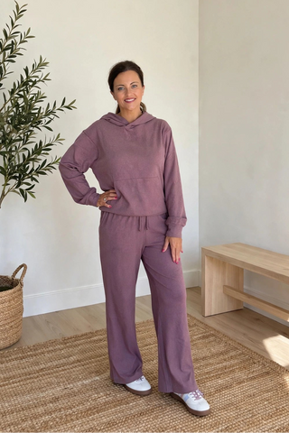 A woman modeling a matching mauve purple lounge set consisting of a long-sleeve hoodie and wide-leg sweatpants, standing in a bright, minimalist room with a potted olive tree and a wooden bench.