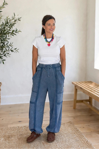 A woman stands in a bright, minimalist room wearing high-waisted wide-leg blue cargo pants, a white t-shirt, and brown shoes. She is positioned next to a potted olive tree and a wooden bench.