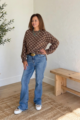 A woman stands in a bright room wearing a brown and beige patterned knit sweater, blue jeans, and white sneakers. She is positioned next to a potted olive tree and a wooden bench on a woven rug.
