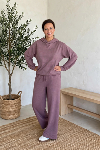 A woman modeling a matching dusty purple lounge set consisting of a cropped hoodie and wide-leg sweatpants, standing in a bright room with a potted olive tree and a wooden bench.