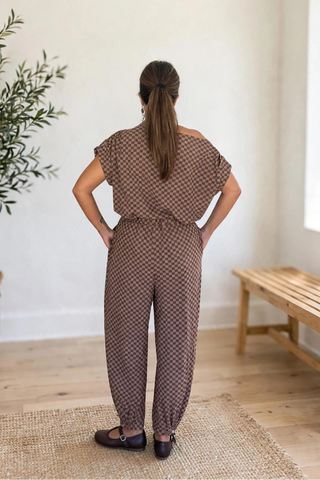 A woman viewed from behind wearing a brown and beige patterned off-the-shoulder jumpsuit in a bright room with a potted plant, woven rug, and wooden bench.