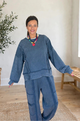A woman wearing a matching dusty blue two-piece lounge set consisting of a long-sleeve top and wide-leg pants, standing in a bright room with a potted olive tree and a wooden bench.