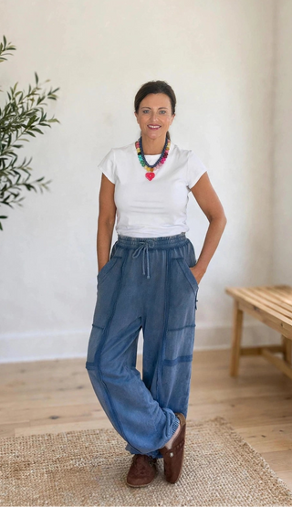 A woman stands in a bright, minimalist room wearing a white t-shirt, blue wide-leg cargo pants, and brown shoes. She is accessorized with a colorful beaded necklace.