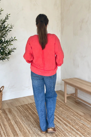 A woman viewed from behind wearing a coral pink long-sleeve top and blue jeans, standing in a minimalist room with a potted olive tree, a wooden bench, and a woven rug.